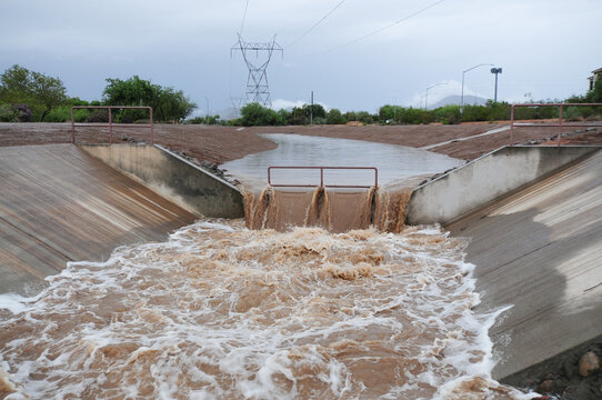 Rainwater runoff flows through a concrete stormwater drainage canal, featuring a central grate and turbulent muddy water under an overcast sky with vegetation and power lines nearby