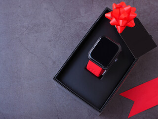 Smartwatch in the black gift box tied with a red bow and black greeting cards on dark grey background. Top view, flat lay with copy space. The concept for the season of giving..