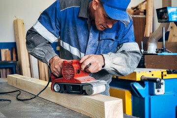 Carpenter in work clothes processes wood with grinder. Polishing of wood surface. Middle-aged man...
