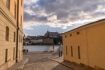 Cobble street and old houses, pier with tourists at the bay Riddarfjärden a winter day with dark clouds and low sun in Stockholm