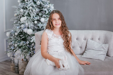 A cute girl with long wavy hair in a white dress on the background of a Christmas tree with a rabbit in her hands.