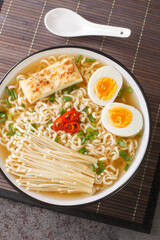Ramen soup with tofu cheese, enoki mushrooms, noodles, green onions and chili peppers close-up in a bowl on the table. Vertical top view from above