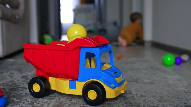 Bright toy lorry filled with plastic balls. Little kid in orange sport suit crawls out of the room. Blurred backdrop.