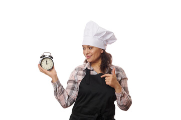 Happy waitress, pastry chef, baker confectioner or new catering establishment owner, in black apron points at black alarm clock showing the opening and closing time of cafeteria, on white background
