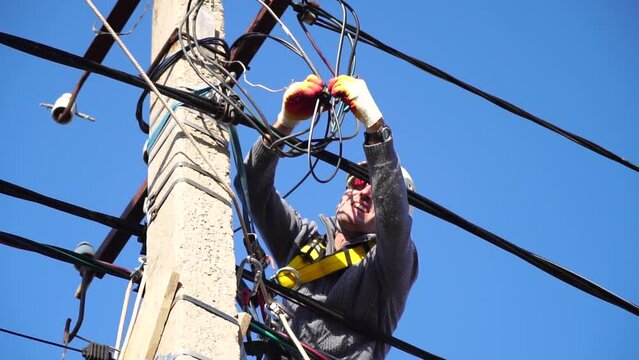 Skilled electrician in helmet fixes wires standing on ladder near high pole against blue sky on summer day backside view. Electrical service and mounting on the pole. Slow motion
