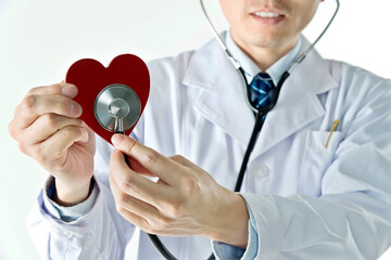 Male doctor with stethoscope examining red heart against white background