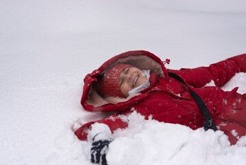 A girl in a red hat and a red jacket in the snow. The girl smiles and lies on her back in the snow. Winter fun. - 552772701
