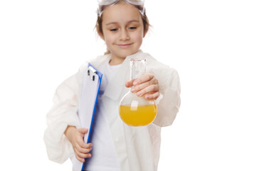 Selective focus on laboratory glass flask with yellow fluid chemical solution, in the hands of blurred adorable school child in lab coat with clipboard, isolated on white background. Chemistry lesson