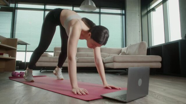 Young Sporty Asian Woman in sportswear watching online video with fitness exercises on laptop. Female fitness practicing leg stretching exercises on yoga mat at home. Pretty lady burning calories