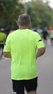 Vertical Screen: Slow motion senior man with gray hair wearing yellow t-shirt running city marathon, other contestants ahead of him. Following shot athletic people taking part in sports event