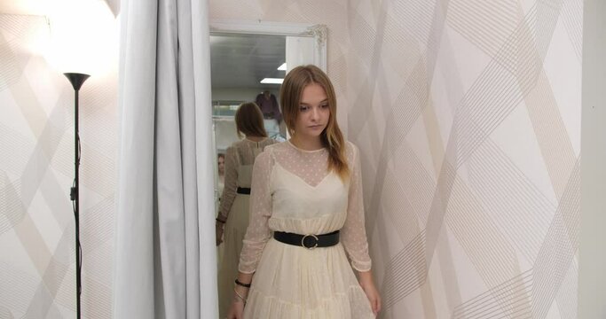 A stylish Caucasian girl is trying on a beautiful dress near the mirror in the dressing room of a clothing store.