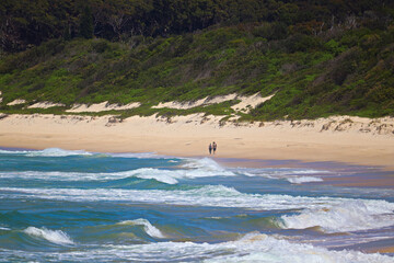 Waves crashing at Fingal Beach with couple walking along the sand in the distance on a sunny day.