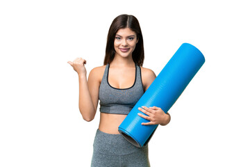 Young sport woman going to yoga classes while holding a mat over isolated chroma key background pointing to the side to present a product