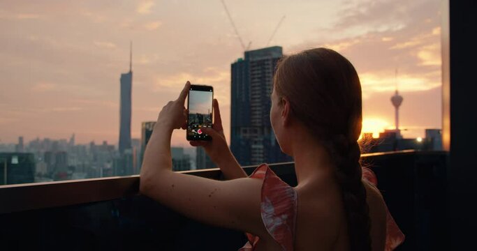 Attractive woman holds a phone in hands and filming a video of evening sunset modern city. Outdoors on terrace with high buildings. Go everywhere traveling concept. Close-up footage.
