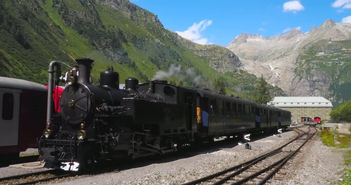 Gletsch, Switzerland - July 15, 2022: A historic train with a smoking steam locomotive on the Furka Railway under the Rhone Glacier