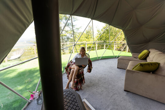 Woman working on laptop geo dome tents. Green, blue background. Cozy, camping, glamping, holiday, vacation lifestyle concept. Outdoors cabin, scenic background.