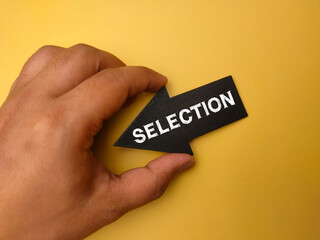 Hand holding arrow wooden board with the word SELECTION on yellow background.