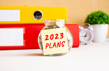 A glass jar with coins is on the work table. There is a sticker on the bank with the text 2023 PLANS.