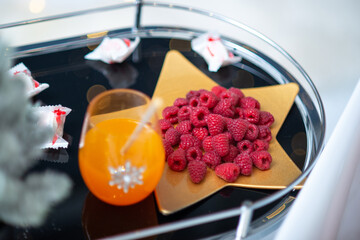 Close-up of small tabel with treats. Raspberry on golden plate in shape of star. Glass of orange juice. Candies. Christmas vibes.