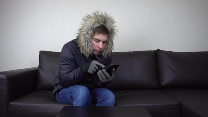 A young man is sitting at home in a winter jacket and trying to keep warm. A man sits without heating and calls on the phone and asks for help.