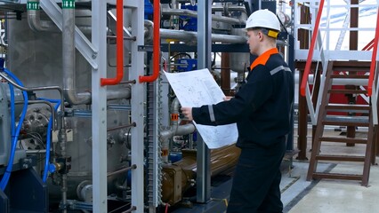 A young engineer at a factory checks the blueprint of a pipeline connection to a compressor unit. An electrical engineer checks the connection of an industrial electric motor according to the scheme.