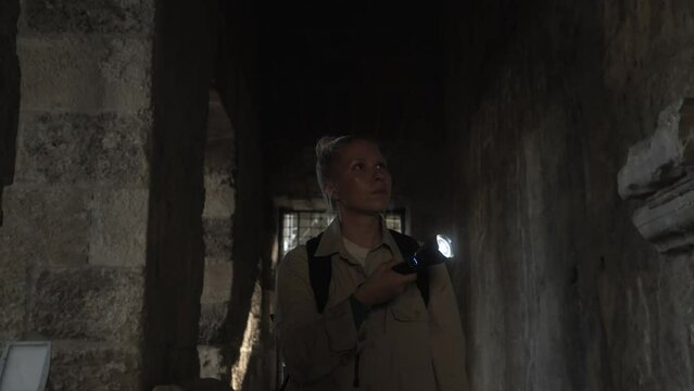 A young female archaeologist stands with a lantern in a dark corridor inside the amphitheater and studies its walls and ceiling. A cinematic frame about ancient excavations. High quality 4k footage