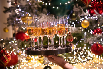 A waiter holding tray with glasses filled with sparkling champagne near decorated christmas tree....