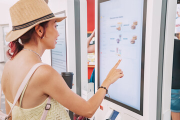 A female customer uses a touchscreen terminal or self-service kiosk to order at a fast food...