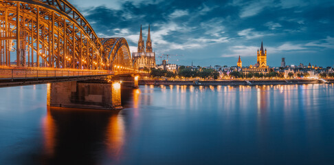 Fototapeta premium Panorama of illuminated bridge over Rhine river whith trains and tourists passing by and the Cologne Cathedral in the evening at the blue hour.