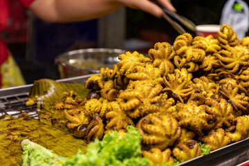 Whole octopus, small size, grilled with sauce until golden. Appetizing. Big piles on banana leaves and blurred lettuce leaves in the foreground. Chef stands blurry in the background. Selective focus.