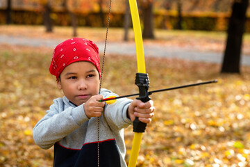 Asian boy in a red bandana aim with a bow outdoor in public park in autumn, close up, selective focus.