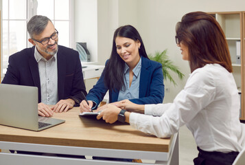 Happy married couple who are clients of bank signs paper document confirming loan for purchase of home. Female bank employee sits at desk in office and shows place for signing contract.