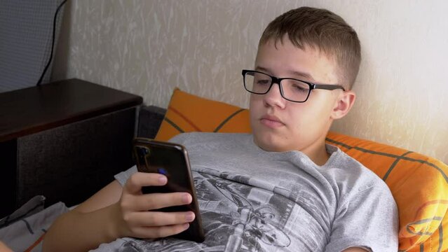 Child in Glasses Lying on a Sofa, Playing Video Games on a Smartphone in Room. Tired cute boy with poor eyesight looks at mobile phone screen before going to sleep. Internet, reading, working online.