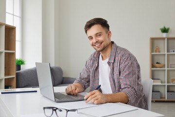 Portrait of happy male student working on his computer. Handsome young man looks at camera and smiles while sitting at table with laptop and writing in notepad. Studying online concept