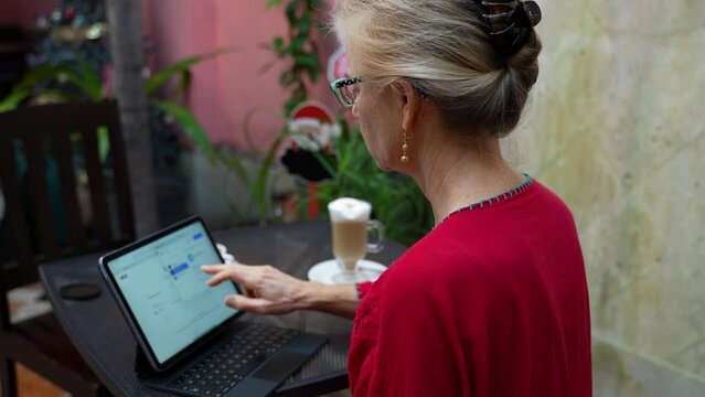 Over The Shoulder View Of Pretty Blonde Mature Woman Working On Tablet Computer At A Cafe With Cappuccino Latte On The Table With Her. Wearing Ethnic Blouse.