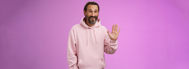 Cheeky charismatic funny happy smiling mature man bearded grey hair in pink hoodie waving palm...