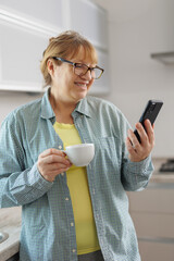 Shot of smiling adult woman using her mobile phone while drinking a cup of coffee in the kitchen at...