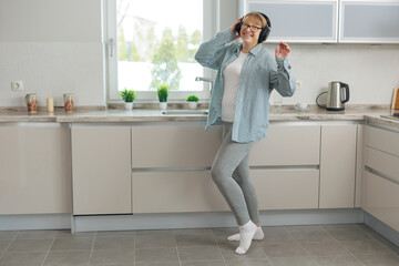 An adult relaxing woman in wireless headphones listens to music and dances in the kitchen. A woman in a blue shirt smiles and dances.