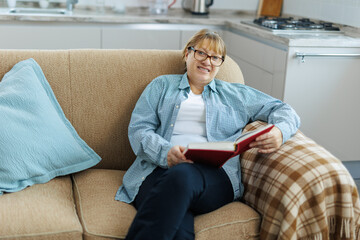 Portrait of mature woman in eyeglasses reading book on sofa at home