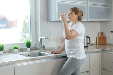 Mature woman in white t-shirt and gray leggings drinks clean water while standing in the kitchen.