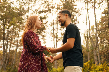 Fototapeta premium Young cute couple are holding hands looking and smiling at each other outdoors in a green park.