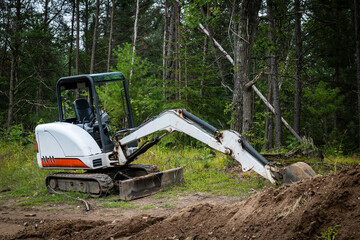 Mini excavator machine with extended arm, at a new home construction site, behind piles of exposed dirt or soil and near forest trees.