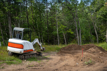 Mini excavator machine at a new home construction site with exposed dirt, a spade, and nearby trees.