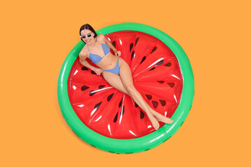 Happy young woman with beautiful suntan and sunglasses on inflatable mattress against orange background, top view