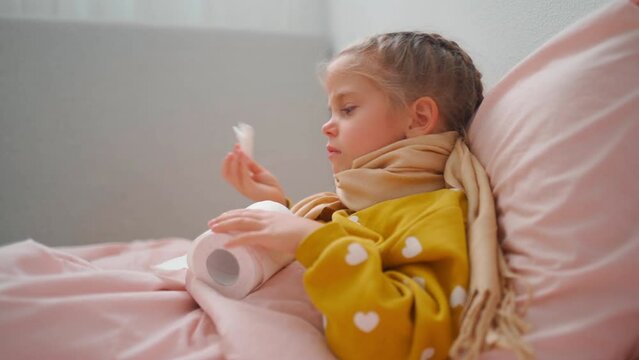 Child girl in plaid and scarf sneezes snot into napkin on sofa at home Child cold flu illness tissue blowing runny nose. toddler girl is lying in bed and blowing nose into tissue paper at home