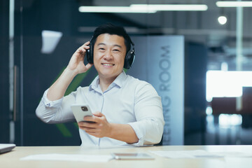 Portrait of a young handsome Asian man in the office. He is sitting at his desk, wearing headphones, holding a phone. He looks at the camera, smiles.