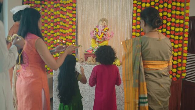 Happy ethnic Hindu Indian family offering a prayer called Aarti to the Ganapati idol decorated with marigold flowers during the Ganesha Chaturthi festival or celebration in an indoor house