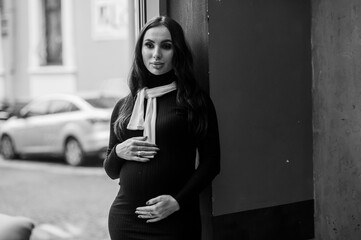 A pregnant woman in a black dress stands near the window