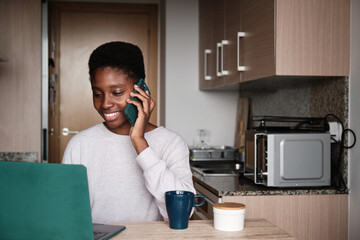 Cheerful black woman speaking on cellphone while using netbook in kitchen