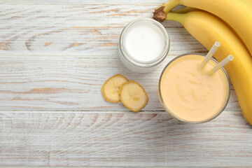 Glass of tasty banana smoothie with straws and ingredients on white wooden table, flat lay. Space for text